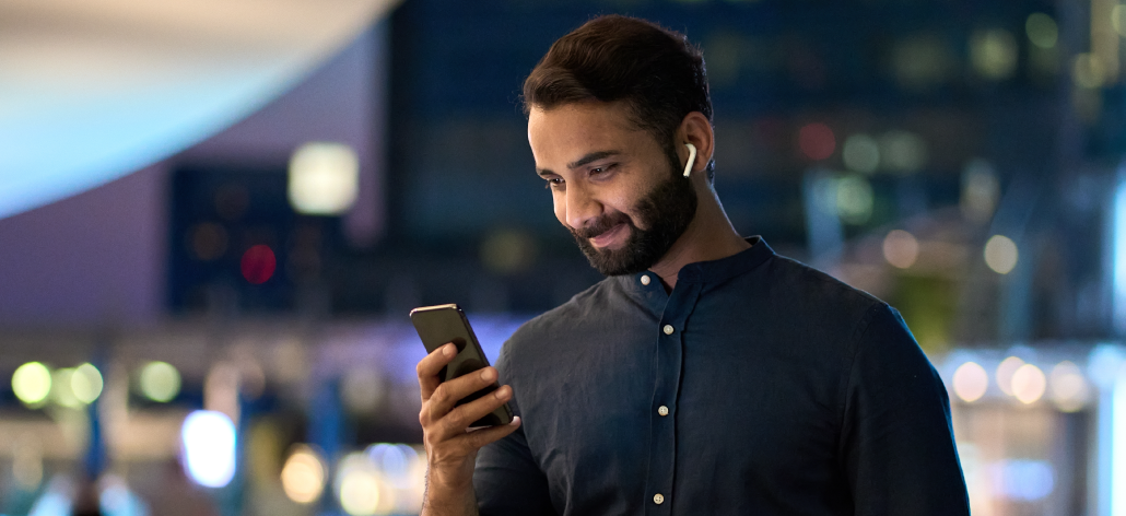 A man smiles at his phone while wearing earbuds.