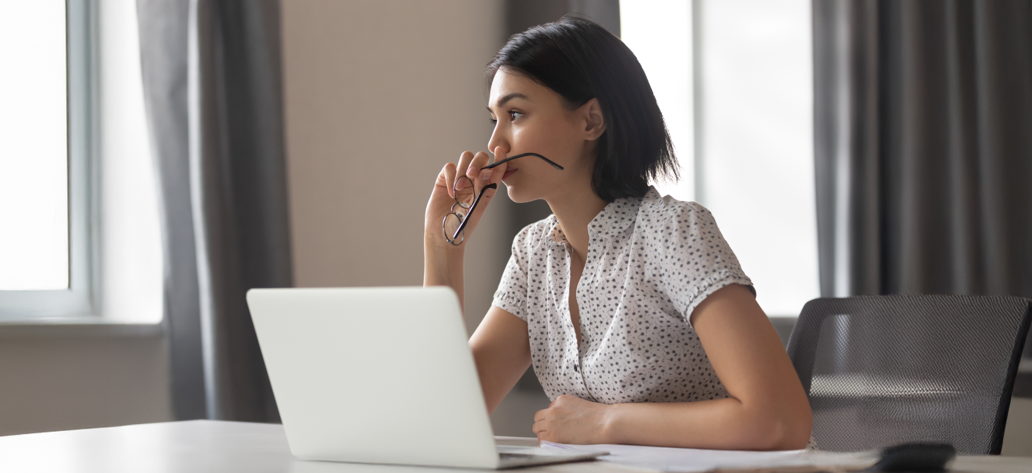 A woman sits at a desk with a laptop holding eyeglasses
