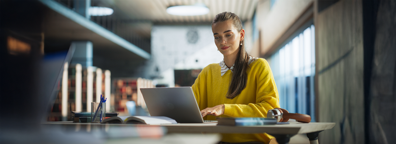 A woman in a yellow sweater works on a laptop at a large desk in a modern library, surrounded by open books and stationery. Natural light fills the space, creating a focused atmosphere.
