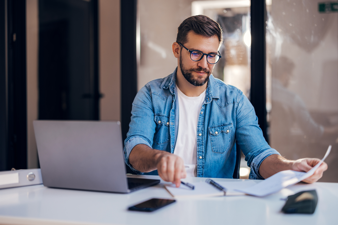 A man wearing glasses and a denim shirt sits at a desk with a laptop, reviewing papers and making notes in a notebook. A smartphone, pen, and folder are on the desk in front of him.