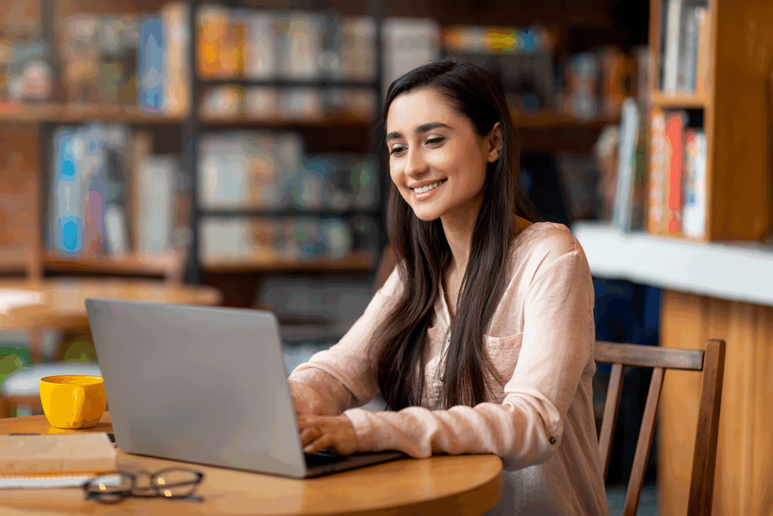 A young woman with long dark hair smiles while working on a laptop at a round wooden table in a cozy library or café. A yellow mug, notebook, and pair of glasses sit on the table beside her.