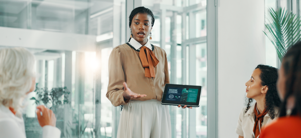 Business woman presenting to meeting table with tablet in hand.